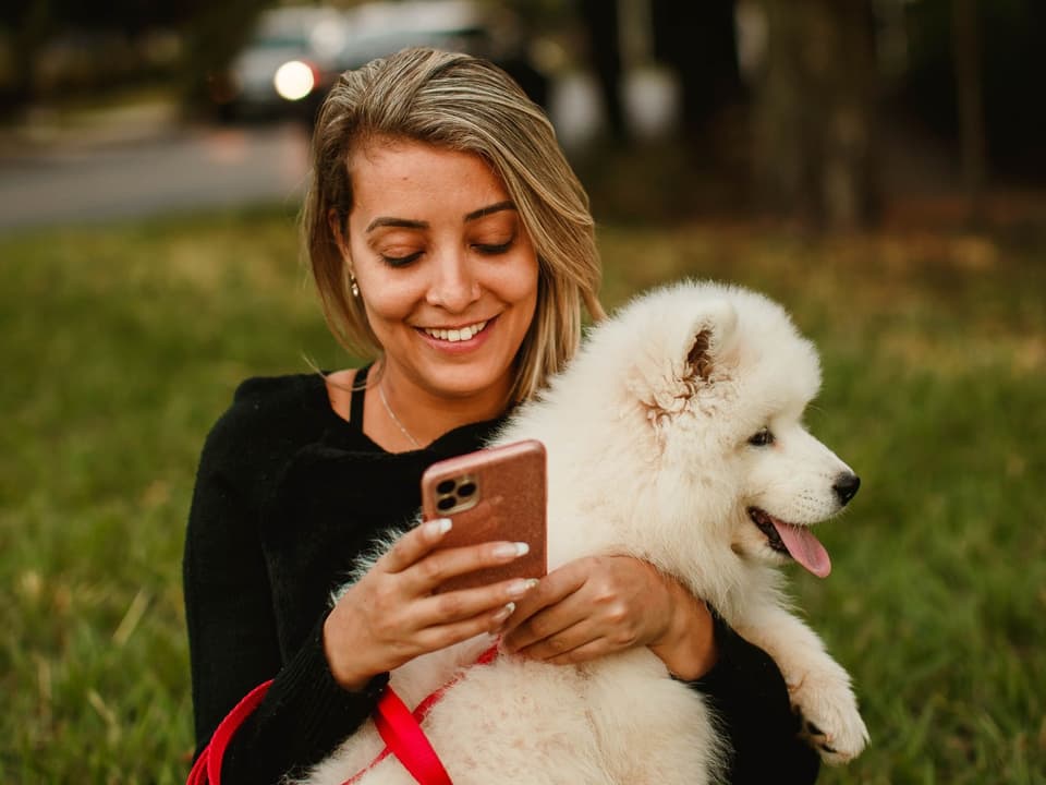 A woman sitting on the grass with her white puppy, smiling as she uses her phone