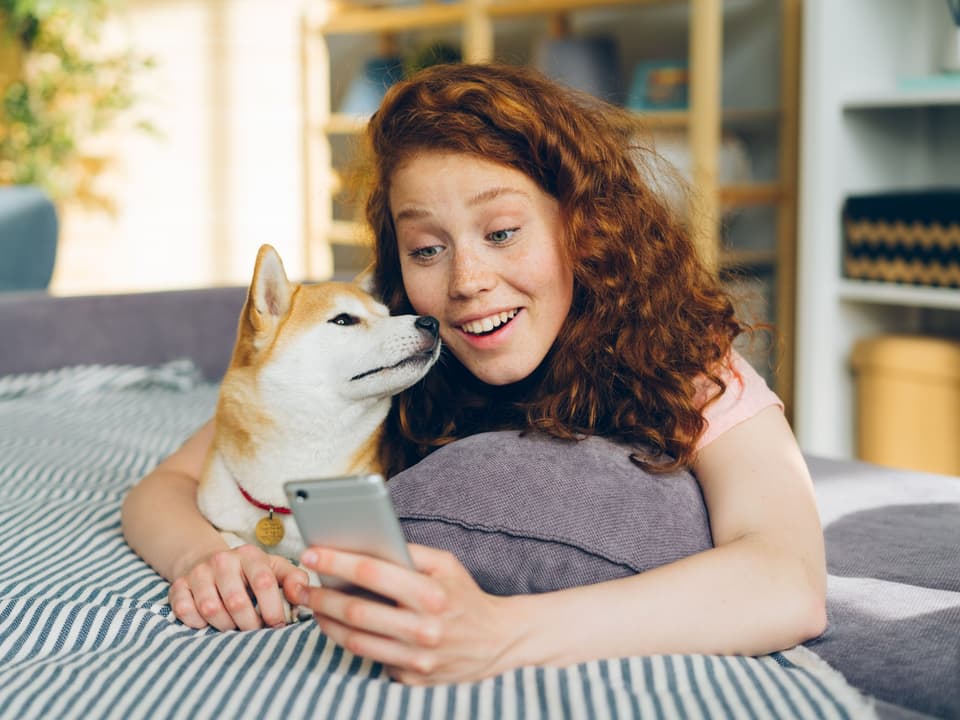 A smiling young woman lying on a sofa beside her dog, looking at her phone
